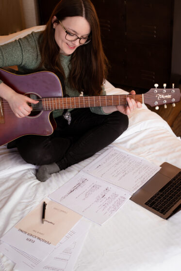 euer-persoenliches-lied-song-schreiben-lassen-hannah-stienen-(c)studiolaecheln-2 Blick schräg von oben: Hannah sitzt auf dem Bett mit ihrer Gitarre. Vor ihr liegt ihr Laptop, ein Fragebogen und Notizen. Sie schreibt gerade ein persönliches Lied für Kunden.