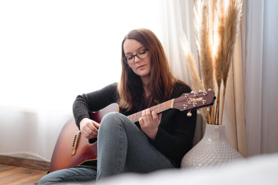euer-persoenliches-lied-song-schreiben-lassen-hannah-stienen-(c)studiolaecheln Hannah Stienen sitzt auf dem Boden mit ihrer Gitarre und probiert Ideen für einen persönlichen Song aus.