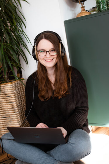 song-schreiben-lassen-euer-persoenlicher-song-hannah-stienen-6 Hannah Stienen sitzt auf dem Boden neben einer Pflanze und vor einer grünen Kommode. Sie hat ihren Laptop auf dem Schoß und Kopfhörer auf. Sie lächelt und hört einen persönlichen Song, den Kunden in Auftrag gegeben und haben schreiben lassen.