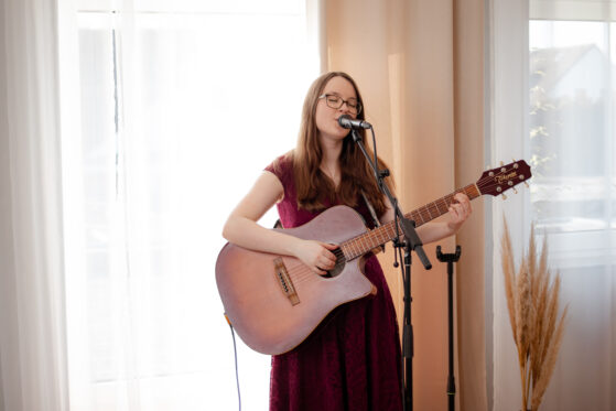 was-kostet-eine-hochzeitssaengerin-hannah-stienen-nrw-(c)studiolaecheln Hochzeitssängerin Hannah Stienen steht vor einem hellen Vorhang und singt.