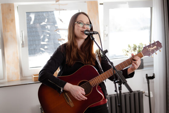 wohnzimmerkonzert-hannah-stienen-2022-(c)studiolaecheln Hannah Stienen bei einem Wohnzimmerkonzert. Sie steht mit Gitarre und Mikrofon vor einer Fensterwand und singt.