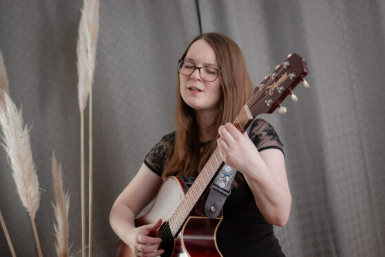 saengerin-beerdigung-trauerfeier-hannah-stienen-2 Hannah Stienen steht als Sängerin zur Beerdigung mit ihrer Gitarre vor einem grauen Vorhang und singt.