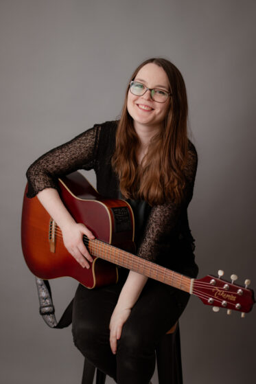 saengerin-fuer-weihnachtsfeier-hannah-stienen-nrw Hannah Stienen sitzt im Fotostudio vor einer grauen Leinwand auf einem Hocker mit ihrer Gitarre und lacht.