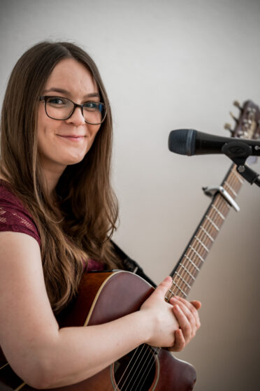 saengerin-firmenevent-hannah-stienen_(c)kroenchens-welt Sängerin Hannah Stienen steht mit ihrer Gitarre am Mikrofon bei einem Firmenevent und schaut in die Kamera.